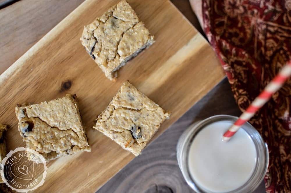 Overhead on three delicious chickpea blondies on a wooden board with a glass of drink and a straw inside of it.