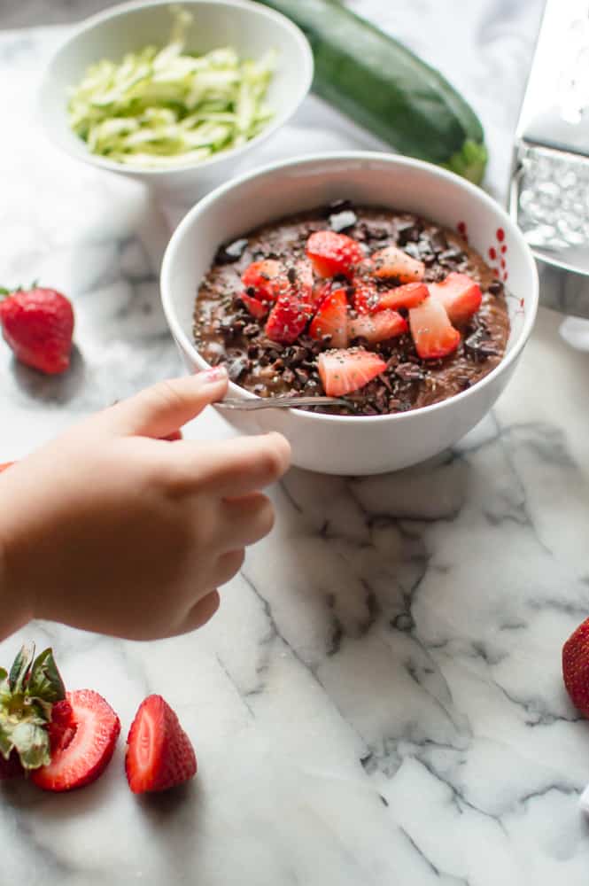A cute little hand reaching for a spoon to try the most delicious Chocolate Zucchini Oatmeal served in a white bowl.