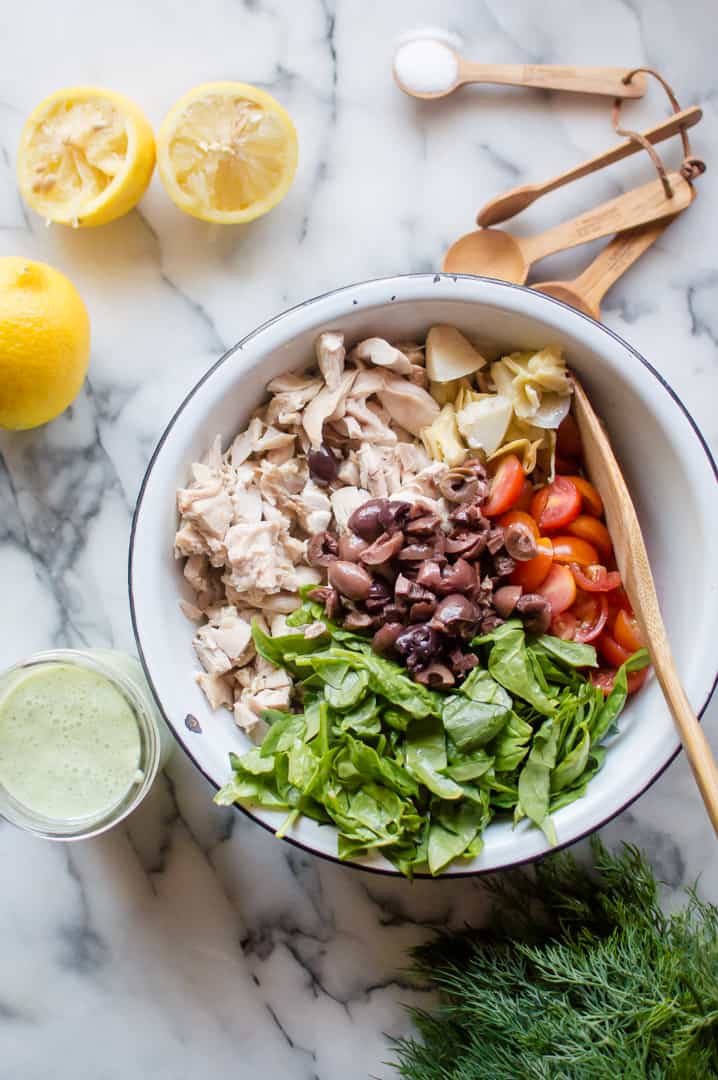 Whole30 Greek Chicken Salad ingredients in a big white bowl with spices in wooden spoons and a couple of lemons on the side