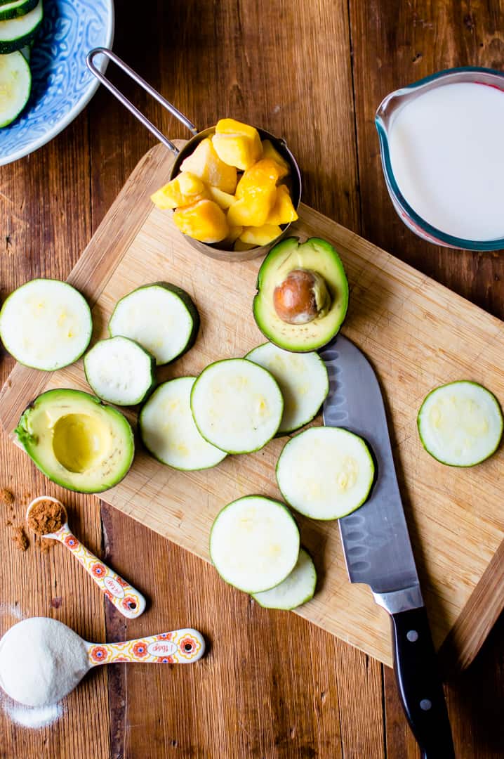 Zucchini slices on a cutting board with a knife and a halved avocado. There is a bowl of mango on the cutting board. 