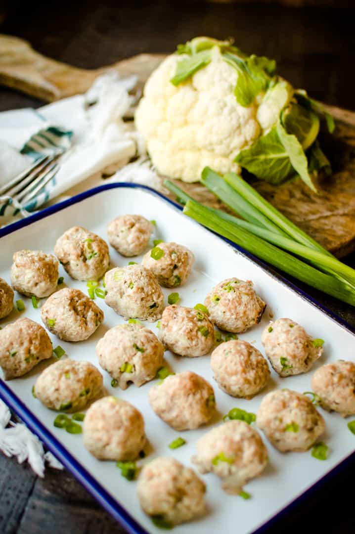 Baked hot chicken cauliflower meatballs straight out of the oven with green onions next to the tray and a head of cauliflower on a wooden table in the background