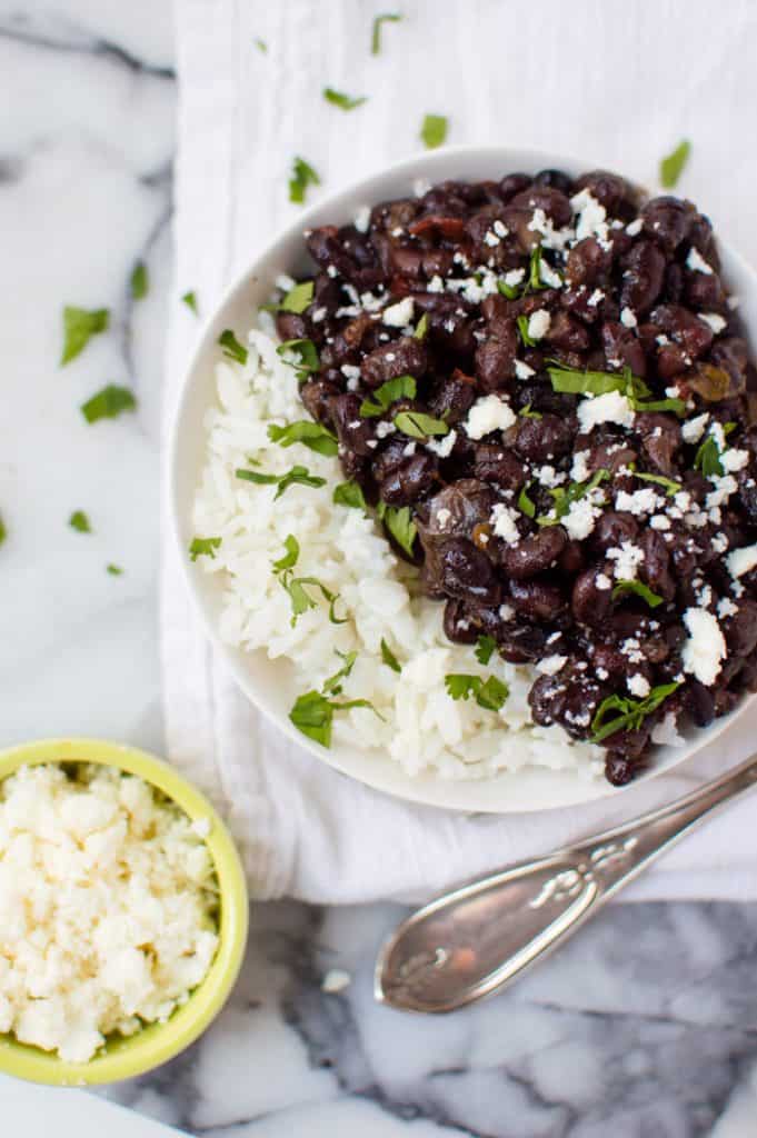 a bowl of finished slow cooker black beans and rice topped with cheese and chopped cilantro on top of a white marble slab and white napkin. 