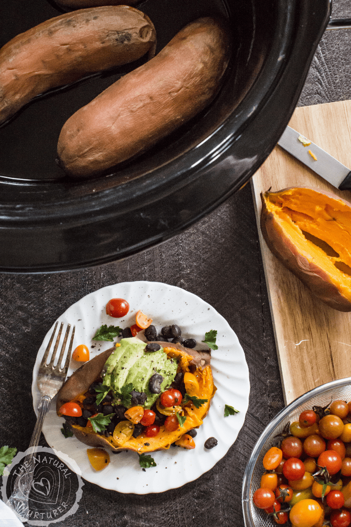 Easy Slow Cooker Sweet Potatoes made in a black slow cooker with a loaded potato served in a white plate with a fork on the side