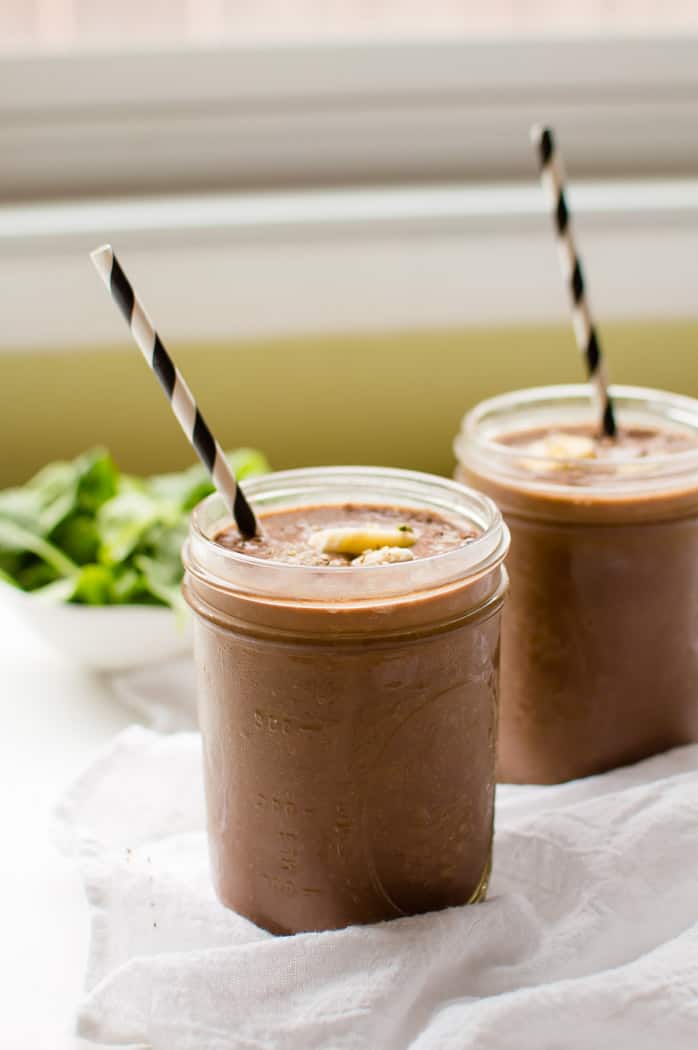 Veggie-loaded Chocolate Banana Smoothie served in two glass jars with two straws on a white piece of fabrics with some greens blurred in the background