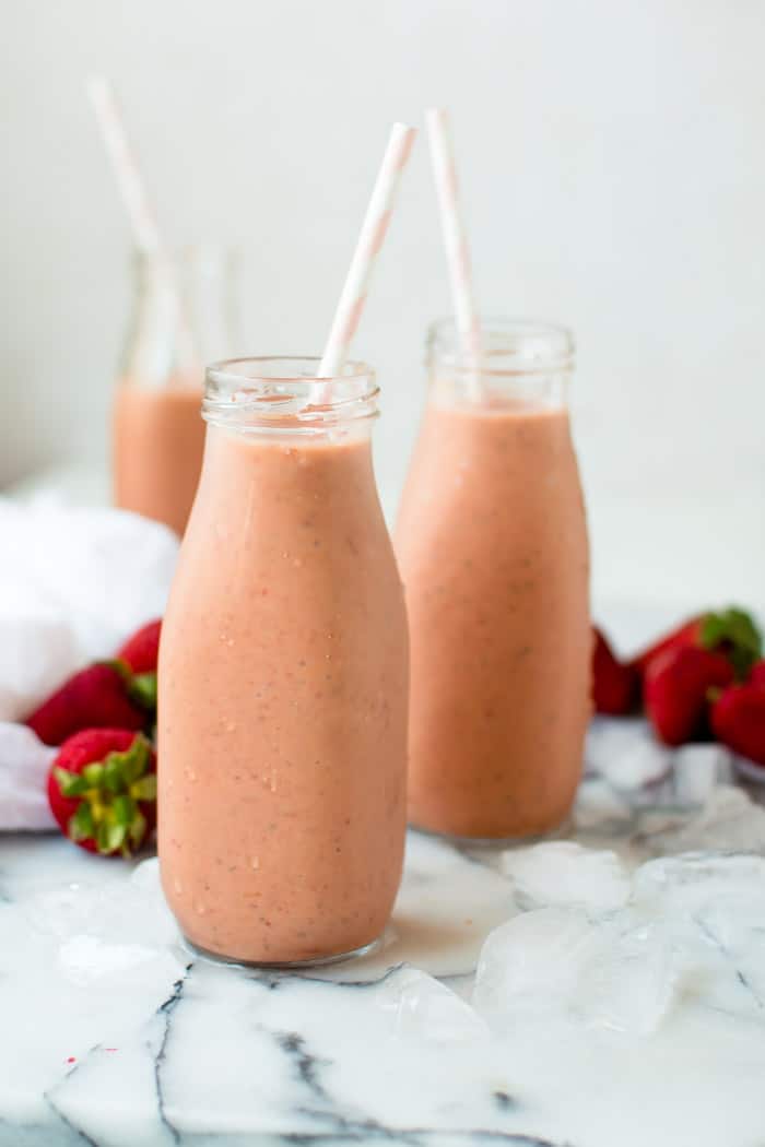 Pink smoothie served in two tall milk jars with white straws inside and strawberries blurred in the background.
