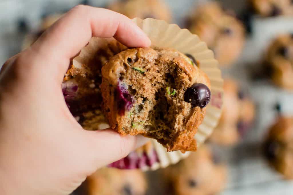 A closeup on the hand holding one of the Almond Butter Blueberry Zucchini Muffins bitten once or twice and revealing the texture inside.