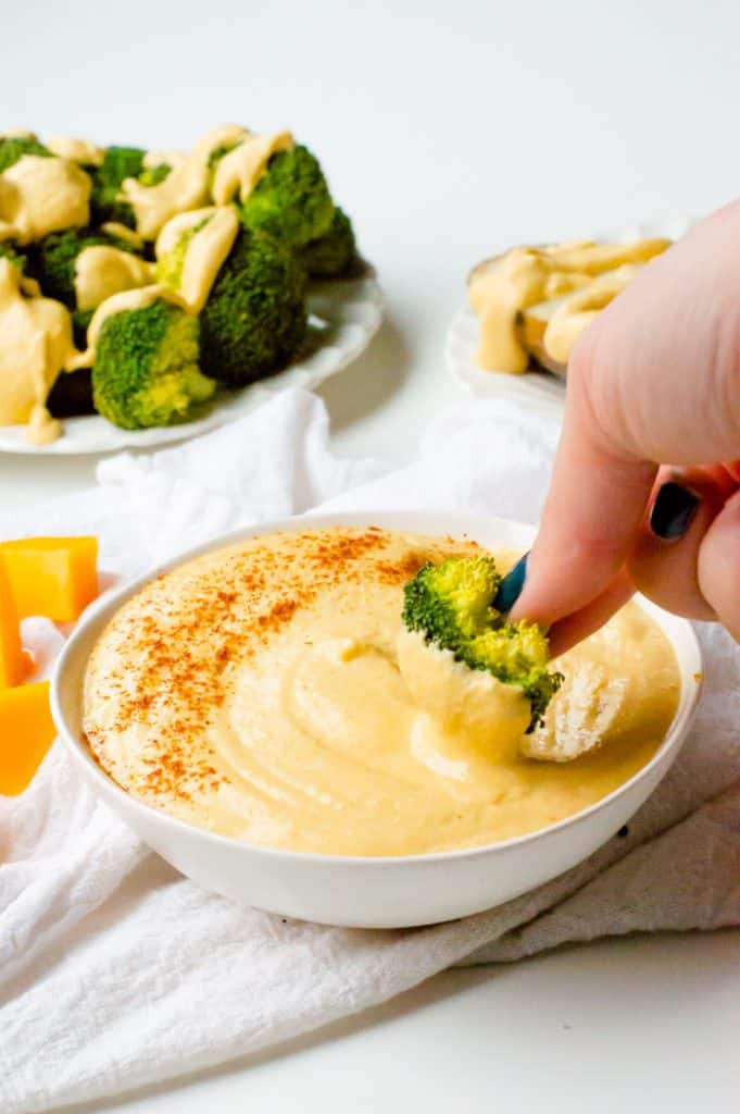 a bowl of vegan cheese sauce with a hand dipping broccoli in it