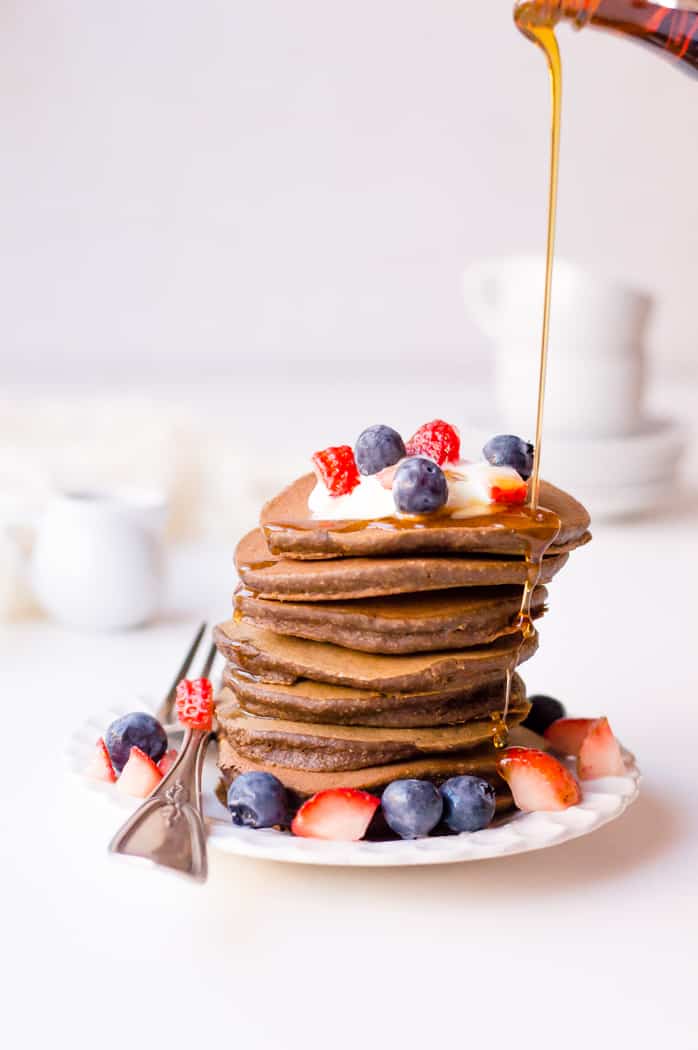 Veggie-packed chocolate pancakes served in a tower with many different berries on top and honey being poured from the top