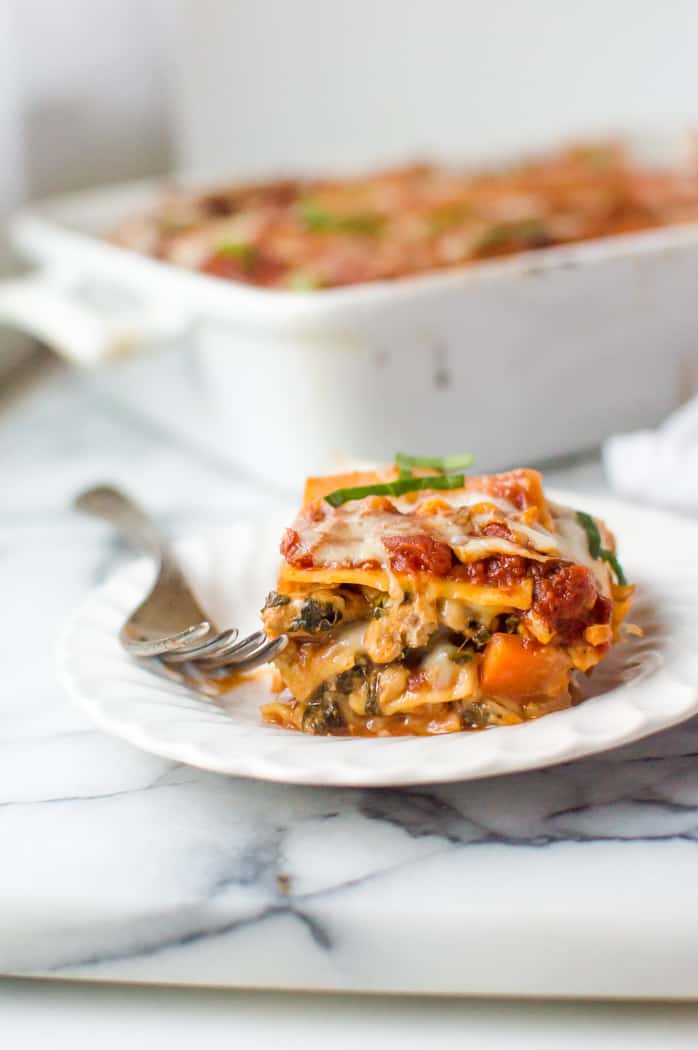 A slice of veggie-loaded turkey lasagna on a plate with a fork. The rest of the lasagna is blurred in the background