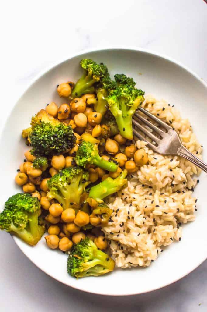 A bowl of broccoli chickpea stir fry with a side of rice and topped with black sesame seeds. A fork, resting on the edge of the bowl, is spearing a floret of broccoli.