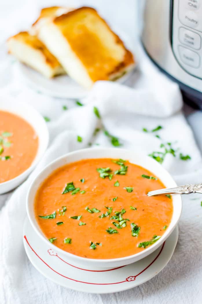 two bowls of veggie-loaded tomato soup on a white marble slab with a white napkin. There is finely chopped spinach sprinkled over the table top and bowls of soup and a grilled cheese sandwich on a plate is sitting in the background.