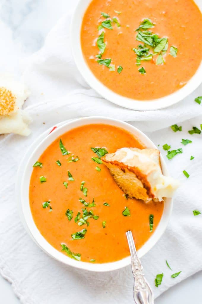 two bowls of veggie-loaded tomato soup on a white marble slab with a white napkin. A half eaten grilled cheese sandwich is dunked in one bowl and there is finely chopped spinach sprinkled over the table top and bowls of soup 