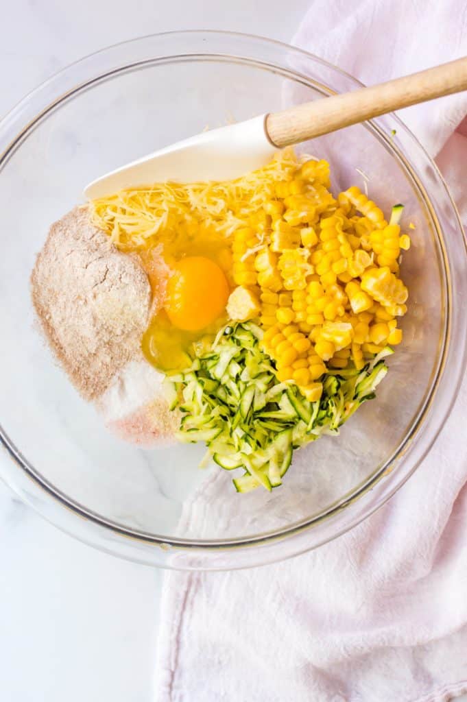 The unmixed ingredients for zucchini and corn fritters in a glass bowl on top of a white table with a white tea towel on the side. There is a spatula rest on the side of the bowl, ready to mix the ingredients. 
