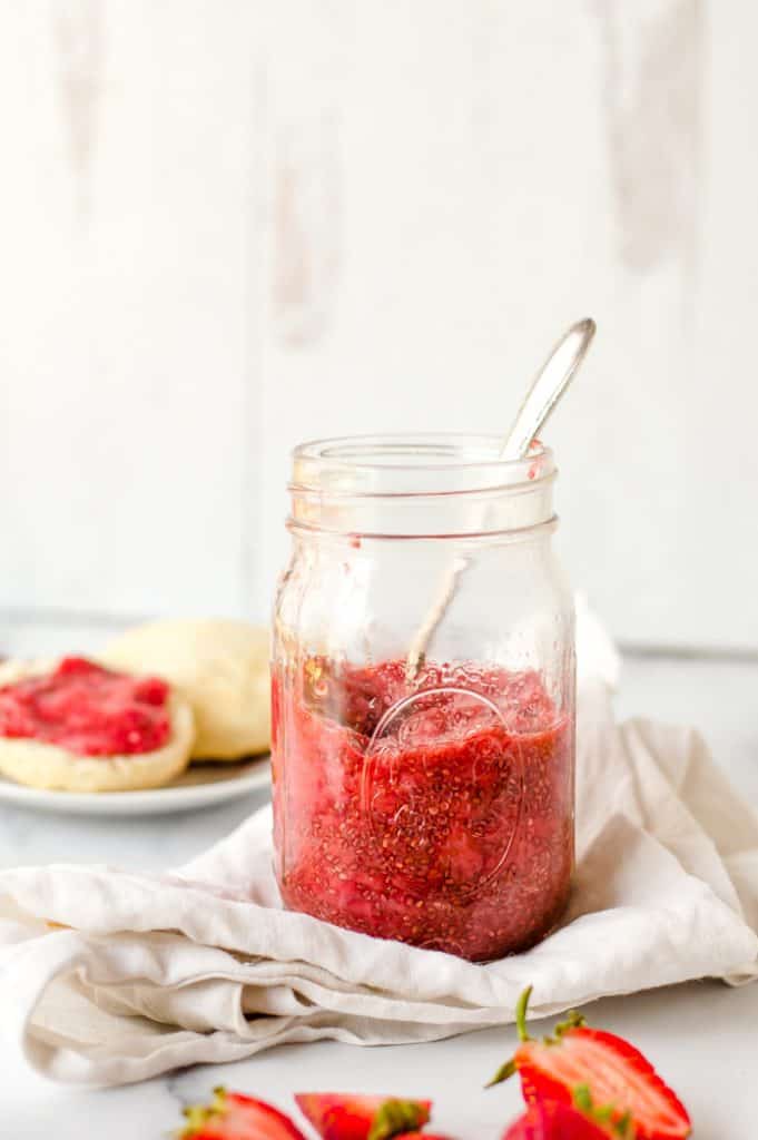 A jar of strawberry chia jam on a counter with a spoon in it and a biscuit with jam on it in the background. 