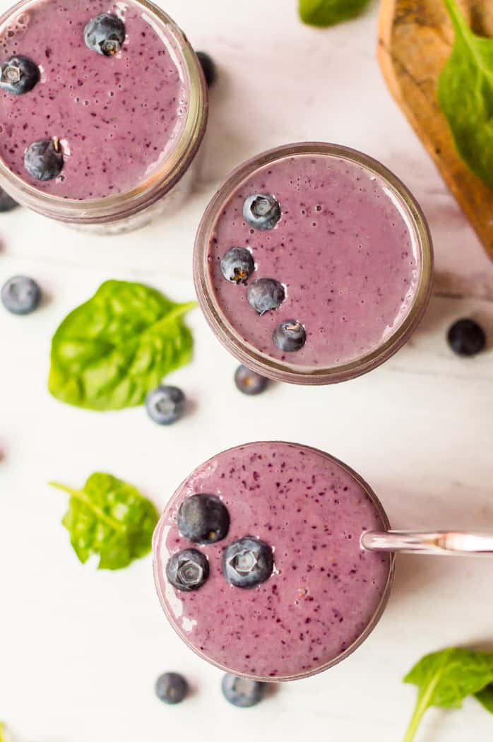 An overhead shot of a blueberry spinach smoothie in 3 glasses topped with fresh blueberries. One glass has a straw in it and there is spinach and blueberries scattered around on the table next to the glasses. 