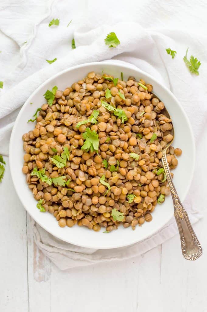 A bowl of cooked brown lentils in a bowl, sprinkled with chopped cilantro.  A spoon is resting on the side of the bowl. 