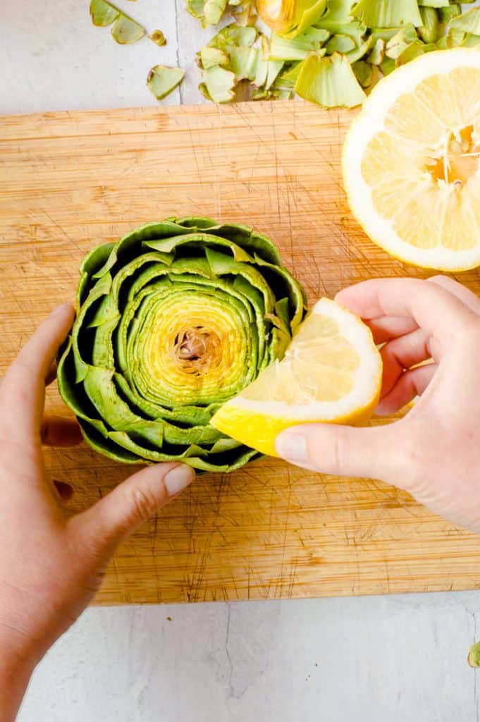 A trimmed artichoke on a cutting board with a hand squeezing a lemon wedge over the top of it. 