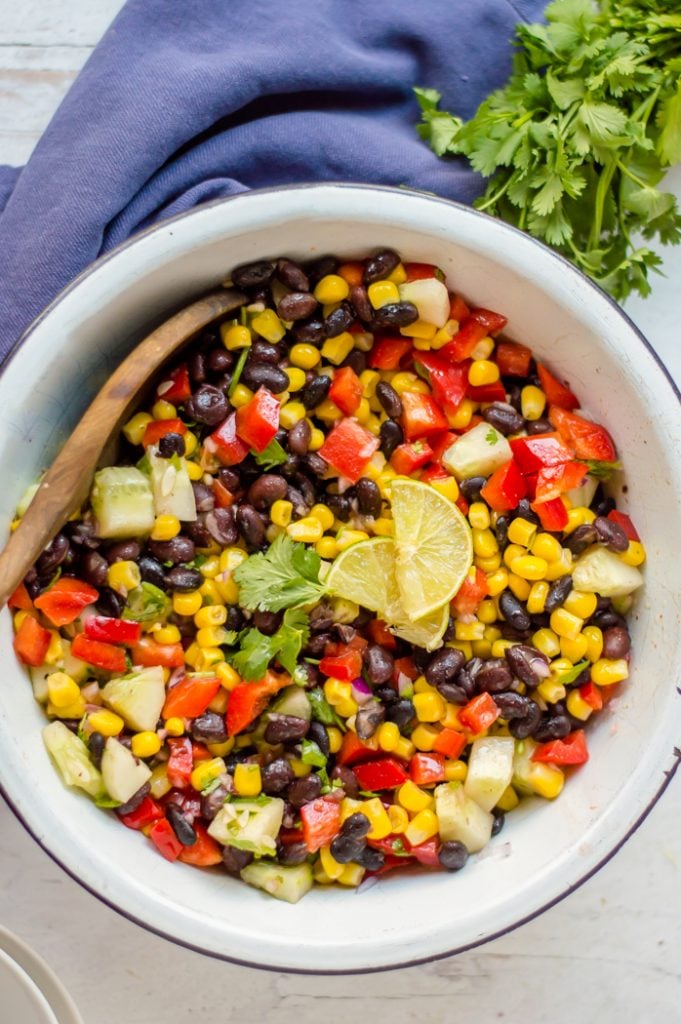 Corn and Black Bean salad in a white bowl with a dark blue napkin under the bowl. There is fresh cilantro on the side and a wooden spoon in the bowl. 