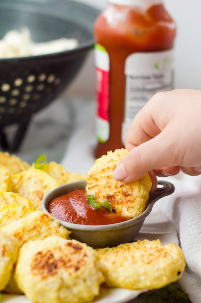 A child's hand taking a cauliflower nugget and dunking it into a cup of ketchup. More nuggets are on the plate around the cup and a bottle of ketchup is in the background with a strainer of cauliflower. 
