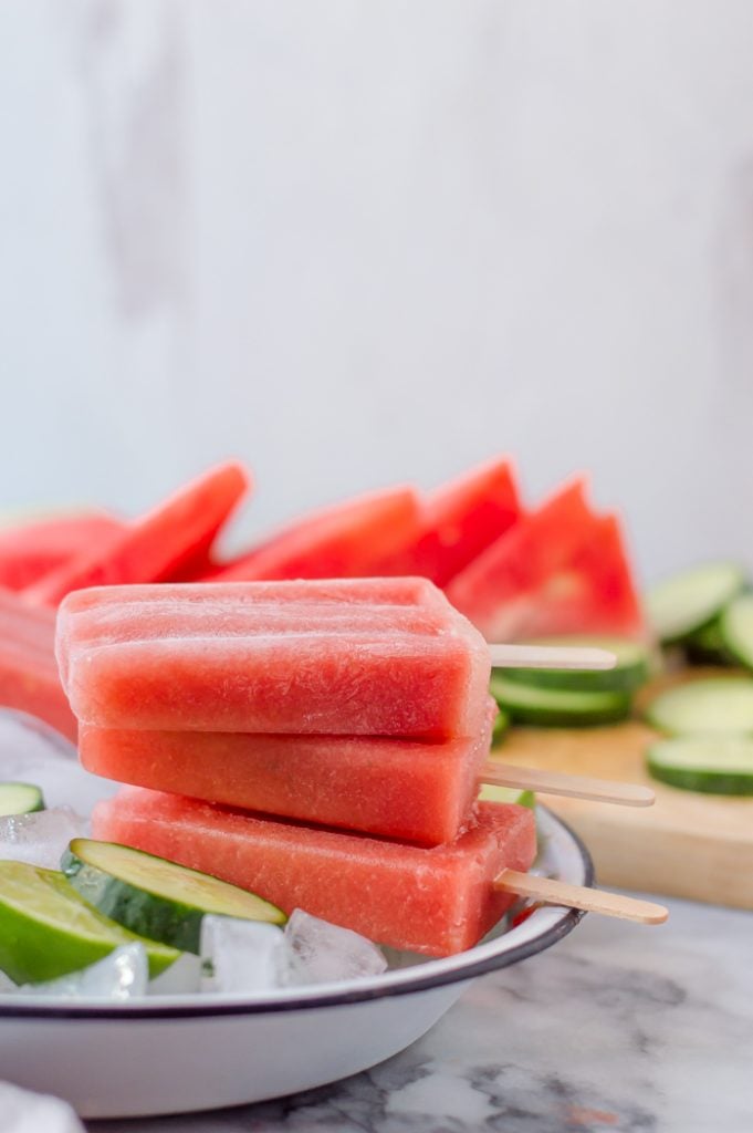 3 watermelon popsicles stacked in a pan of ice with cucumber and watermelon around it. 