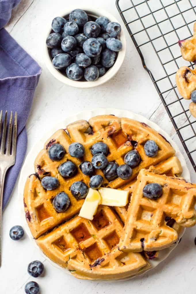 A blueberry zucchini waffle on a plate topped with butter, syrup and fresh blueberries. There is a bowl of blueberries next to the plate and a fork and blue napkin on the side. 