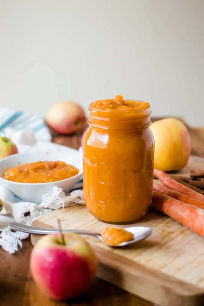 A jar of veggie-loaded homemade applesauce on a cutting board with apples, carrots and a bowl of sauce next to it. 