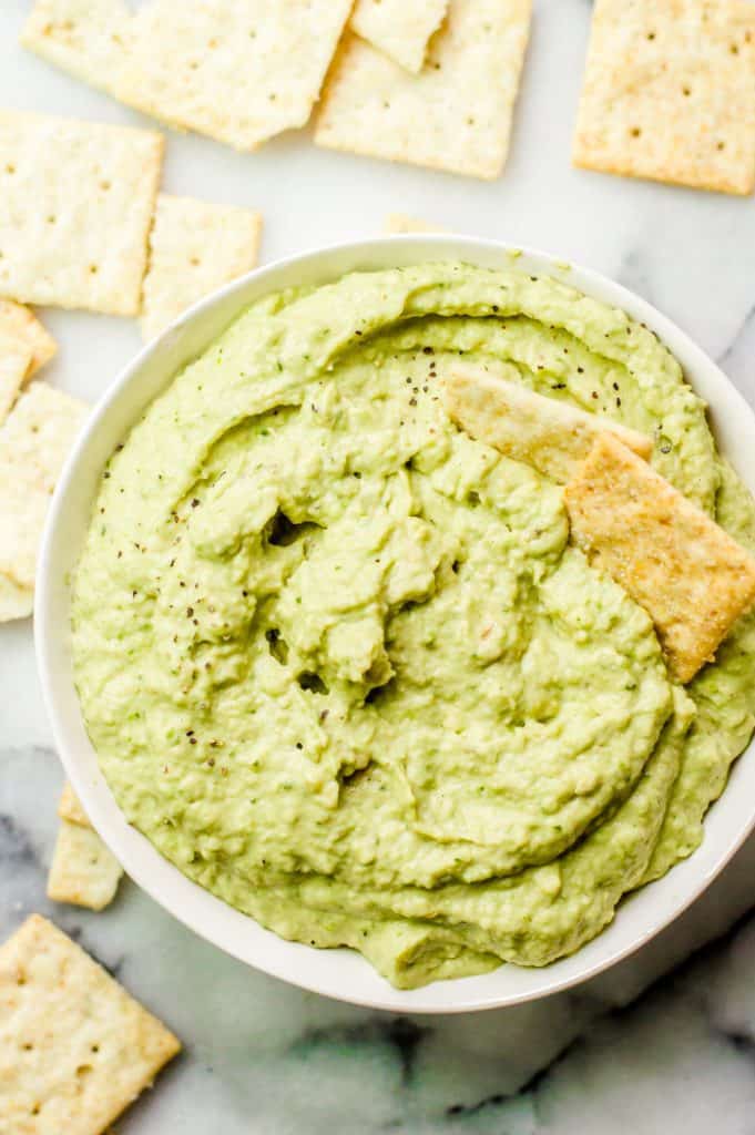 A bowl of zucchini avocado dip with two crackers in it and other crackers scattered around. 