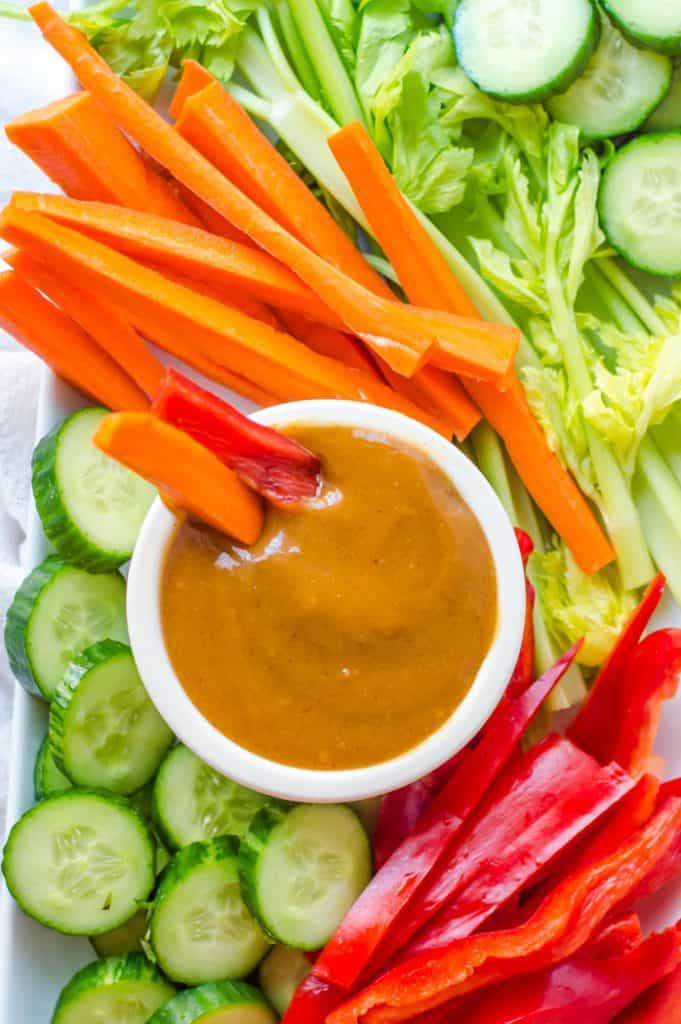 A bowl of peanut sauce on a tray with veggies. A carrot and bell pepper stick are in the bowl of sauce. 