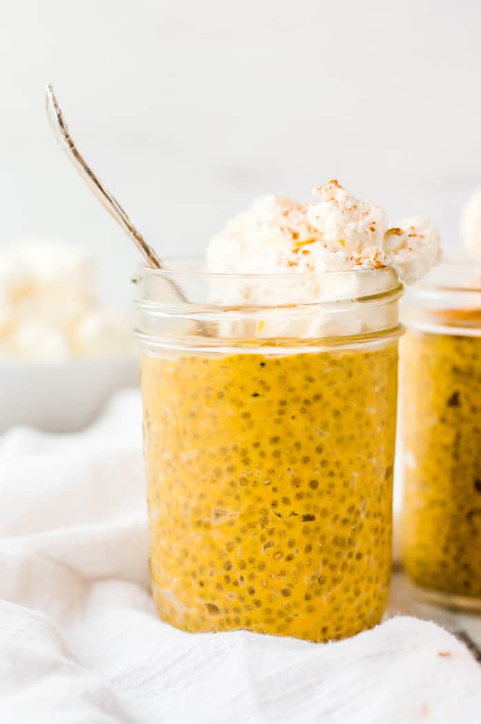 Two jelly jars filled with pumpkin chia seed pudding and topped with whipped cream and cinnamon. One jar has a spoon in it.