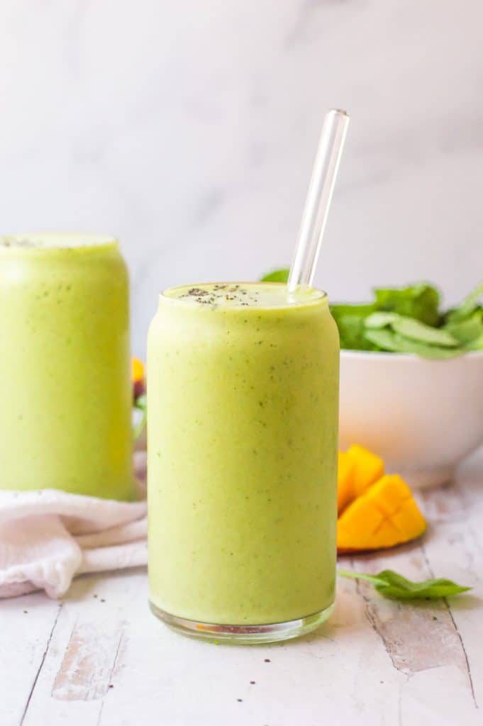 Spinach Mango Smoothie in two glasses with fresh mango and spinach in the background. One glass has a glass straw in it.