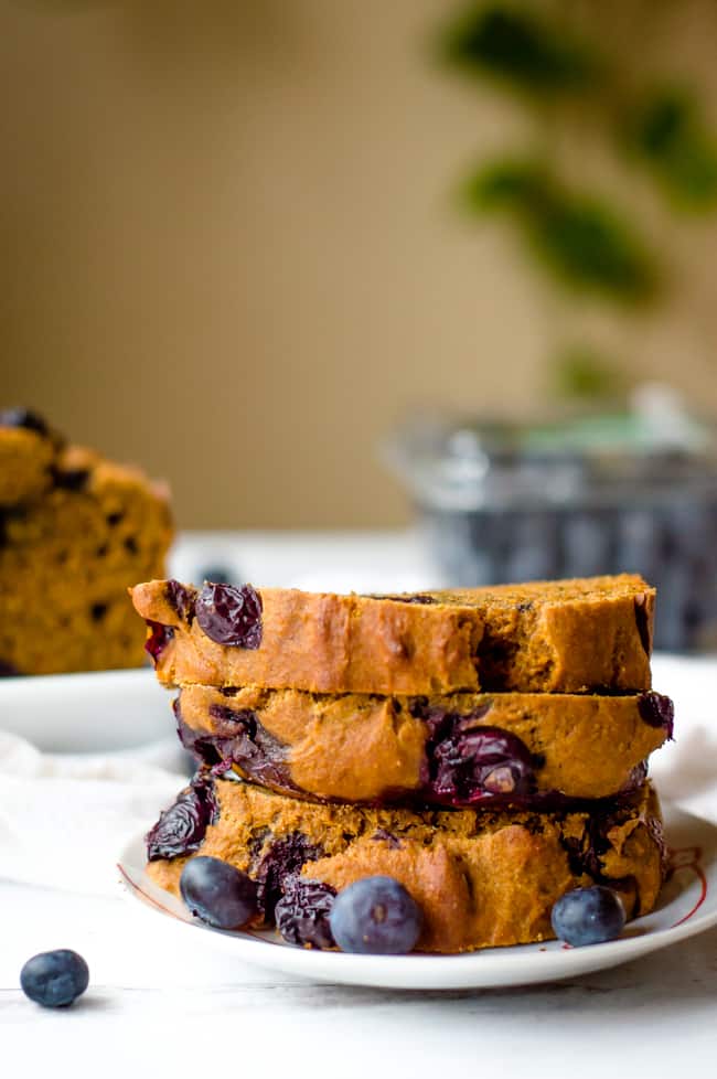3 slices of sweet potato blueberry bread stacked on top of one another with the loaf in the background and a clamshell of blueberries.
