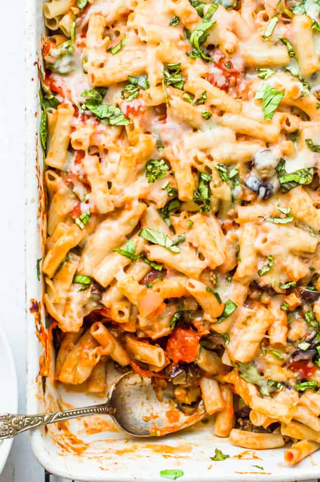 Baked Pasta with Eggplant  in the baking dish topped with chopped spinach and a spoon has takes a serving out of it. 