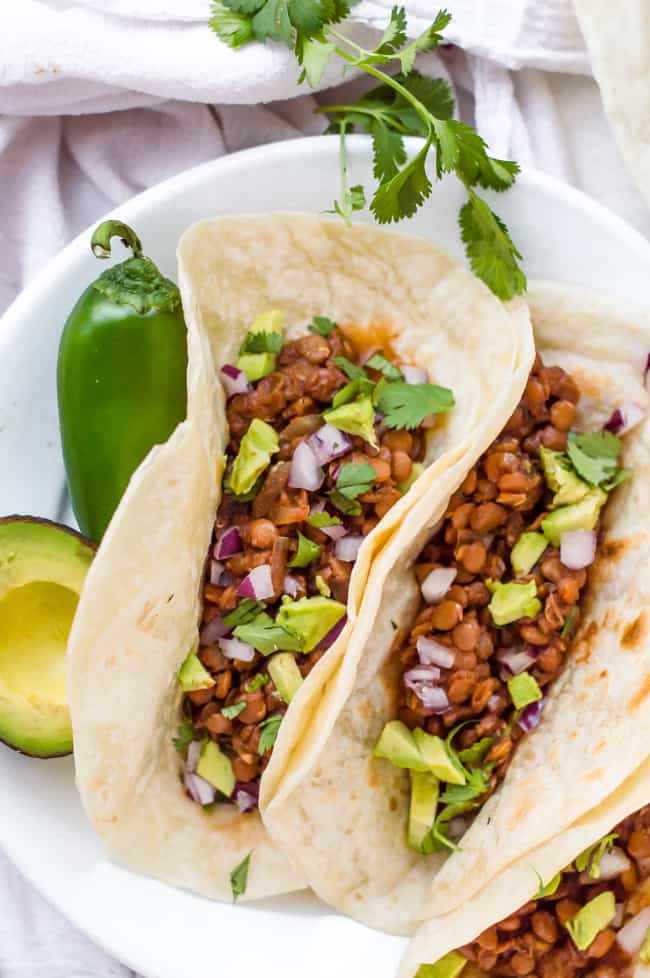 Vegan slow cooker lentil tacos on a plate in tortillas. They have onion, avocado and cilantro sprinkled on top. A jalapeño and avocado are on the plate too. 