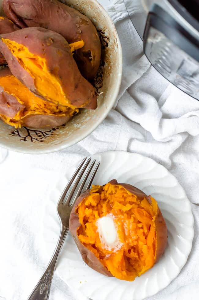 A cooked Instant Pot sweet potato on a plate with a pat of butter and a fork. A bowl of cooked sweet potatoes and an Instant Pot are sitting in the background. 