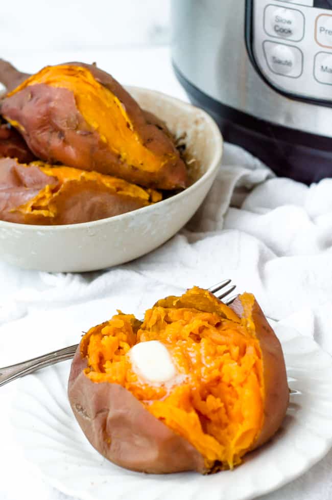 A cooked Instant Pot sweet potato on a plate with a pat of butter and a fork. A bowl of cooked sweet potatoes and an Instant Pot are sitting in the background. 