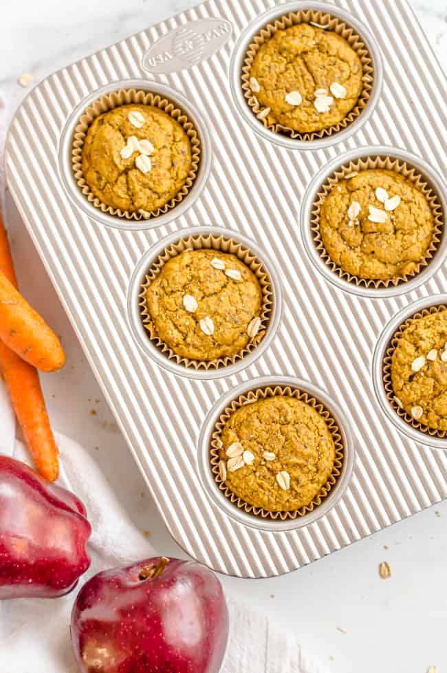 Carrot apple vegan muffins in a muffin tin, right out of the oven. There are apples and carrots next to the pan.