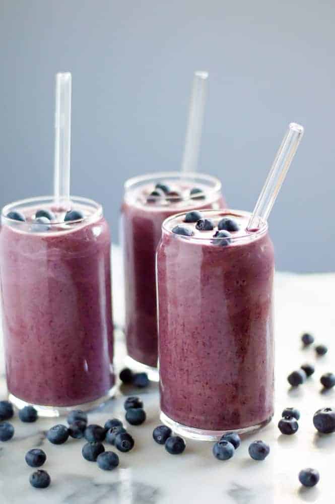 3 cauliflower blueberry smoothies in glasses topped with blueberries and with fresh berries on the table around them. 