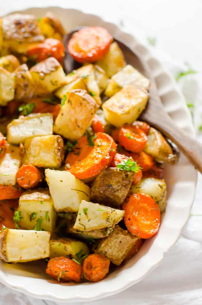 A bowl of roasted carrots and potatoes with fresh parsley sprinkled on top with a wooden spoon on the side. 
