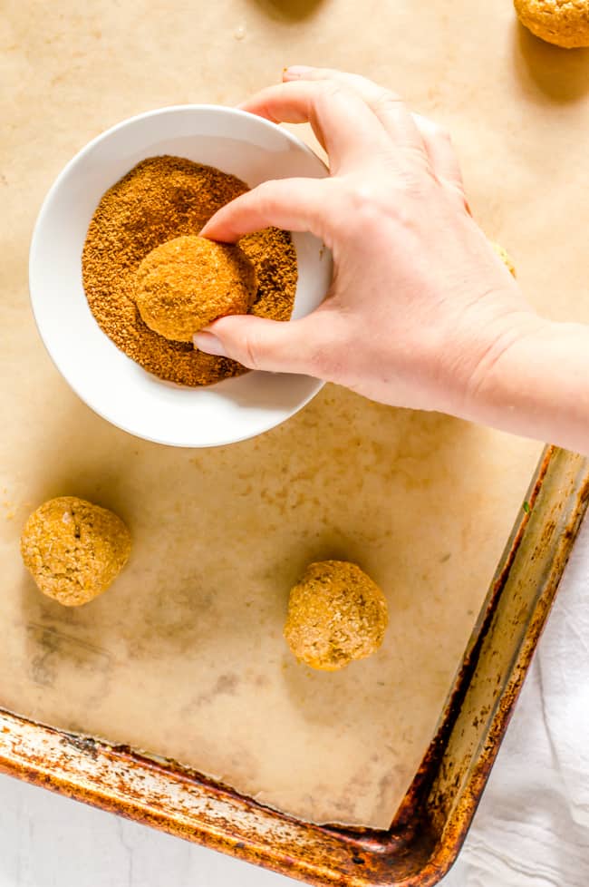 A hand rolling a sweet potato cookie in a bowl of coconut sugar before being baked. The bowl is on a baking sheet with other cookie balls