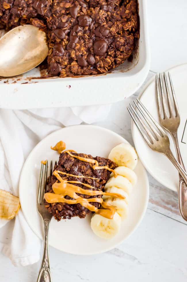 A slice of chocolate baked oatmeal on a plate with peanut butter drizzled over the top, sliced banana next to it and a fork on the plate. The tray of the baked oatmeal is next to the plate and so are extra forks and plates.