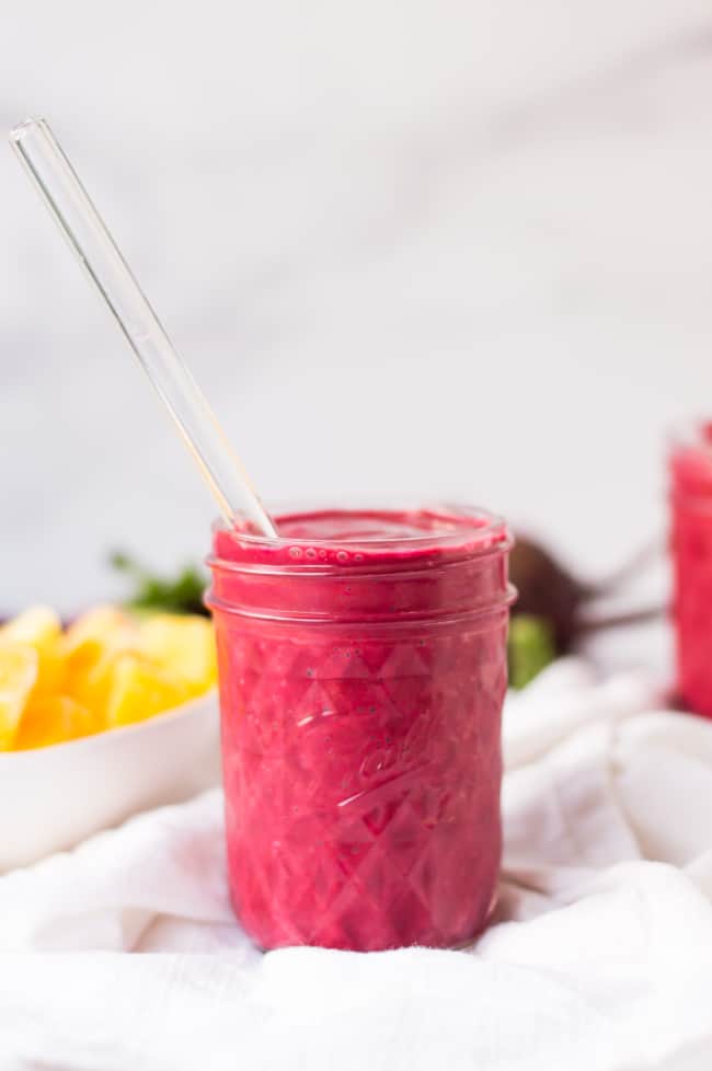 A red velvet smoothie in a jelly jar with a glass straw. There is a white napkin under the jelly jar and a bowl of frozen mangos and fresh beets are in the blurred background.