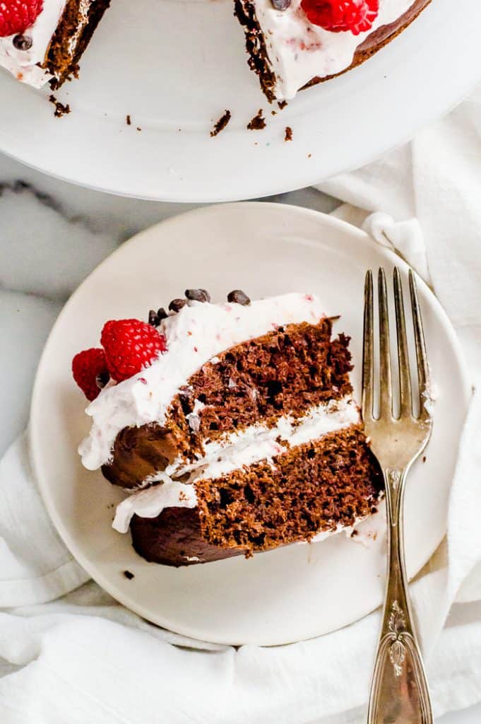 A slice of chocolate beetroot cake on a plate with a fork. It is topped with whipped cream, fresh raspberries and chocolate chips. 