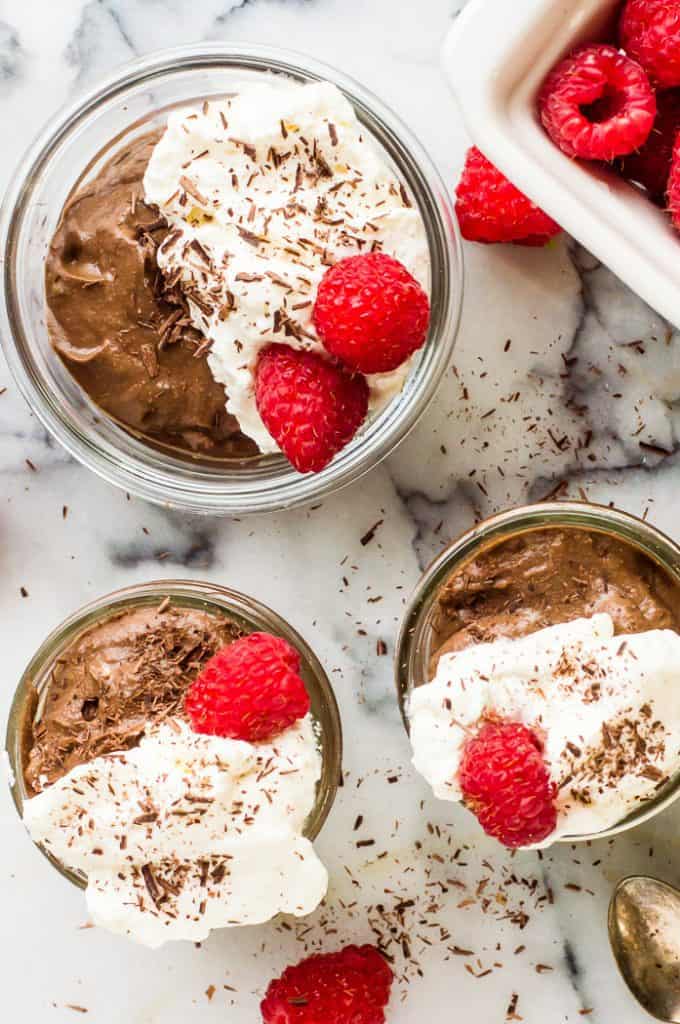 3 cups of chocolate avocado mousse topped with whipped cream, shaved chocolate and a fresh raspberry. There is a container for fresh raspberries next to the the cups.