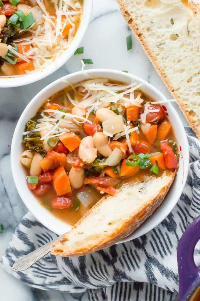 A bowl of kale and white bean soup topped with shredded cheese. A slice of bread is in the bowl with the soup and another bowl and bread are next to the bowl. 