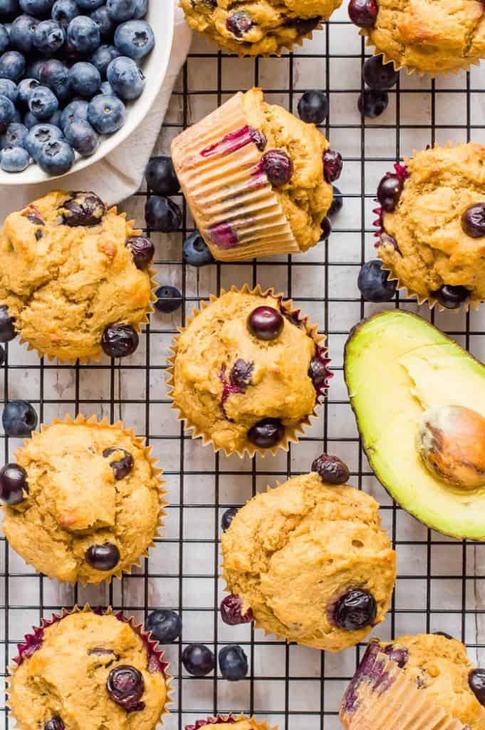 Blueberry avocado muffins on a cooling rack with fresh blueberries and a sliced avocado on the rack.