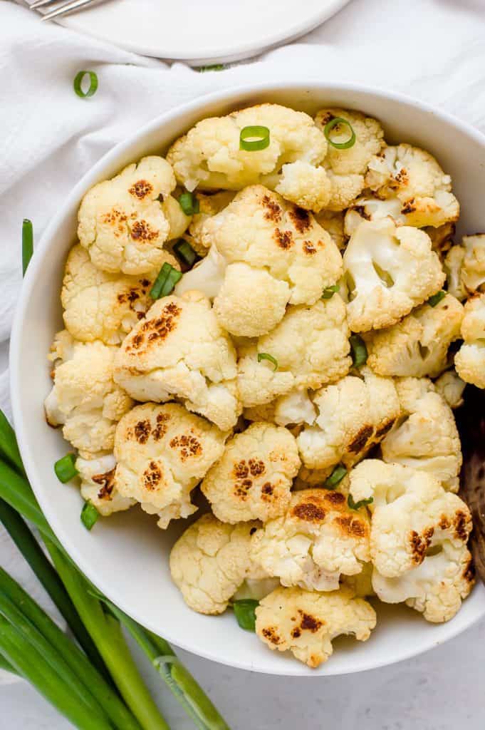 A bowl of roasted cauliflower in a white bowl and topped with sliced green onions. There is a bunch of green onions next to the bowl. 