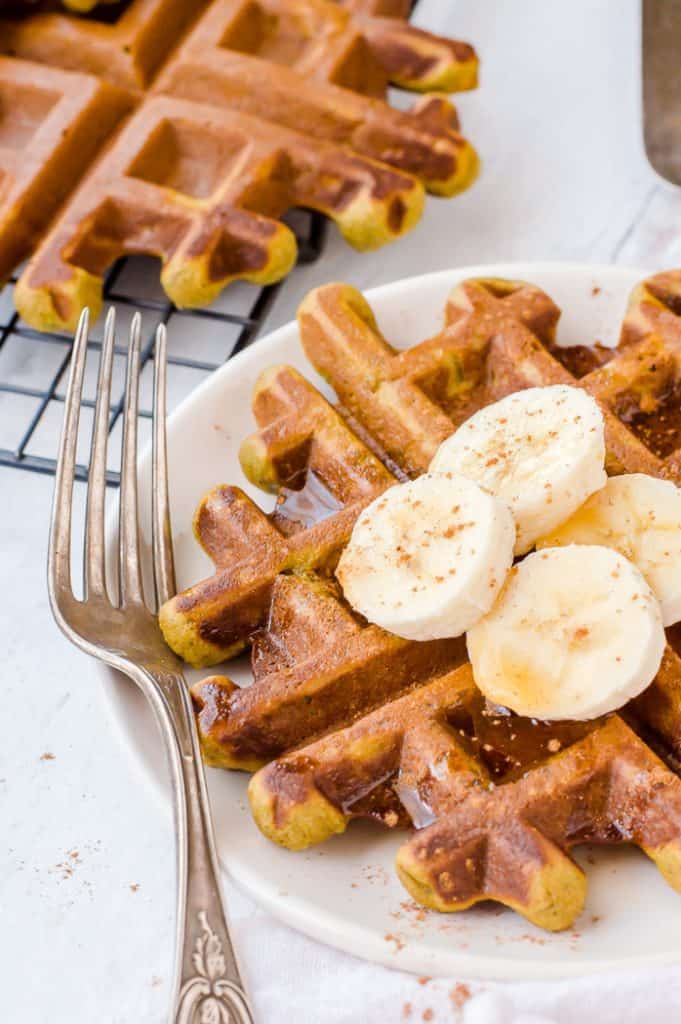 A superhero gluten free waffle with sliced bananas and drizzled maple syrup on top. There is a fork on the plate with the waffle and there are more waffles to the side on a cooling rack.