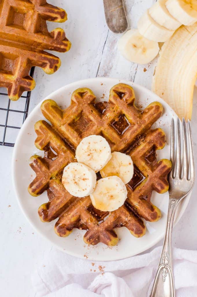 A superhero gluten free waffle with sliced bananas and drizzled maple syrup on top. There is a fork on the plate with the waffle and there are more waffles to the side on a cooling rack.