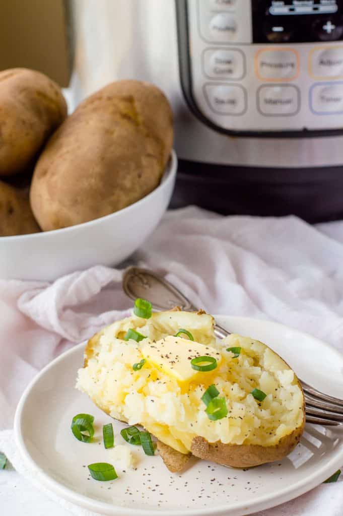 An Instant Pot baked potato on a plate with sliced green onions, pepper and butter. There is a fork on the plate, a bowl of baked potatoes and the pressure cooker in the background.