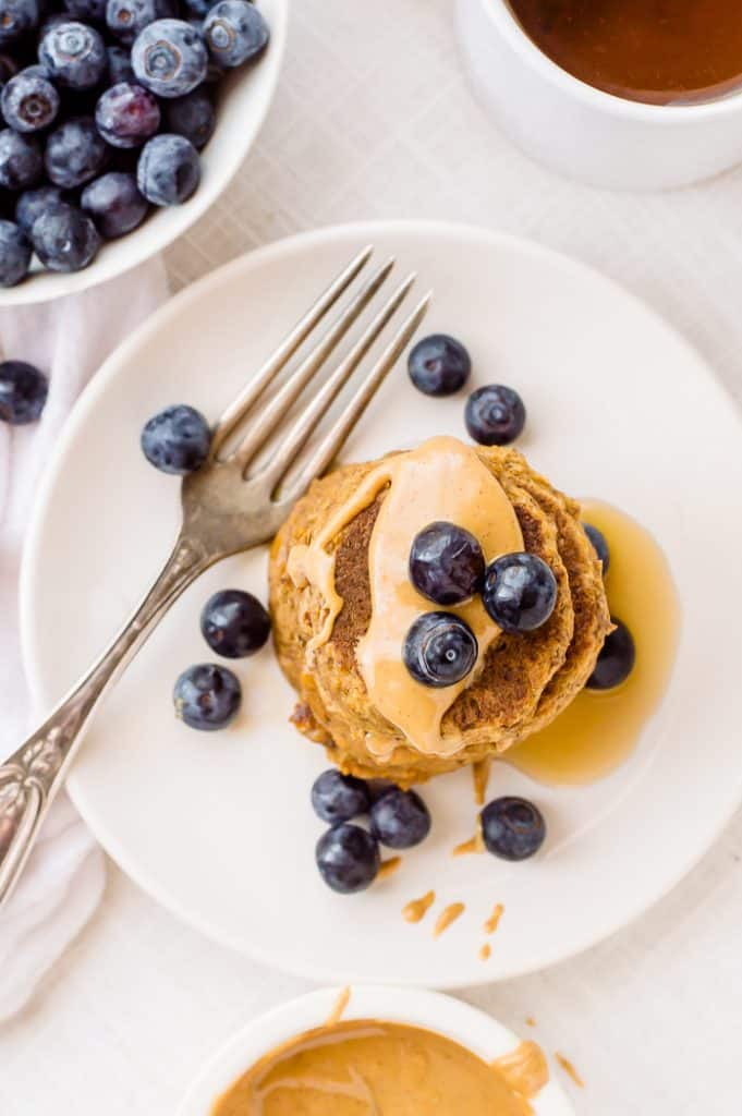 A stack of vegan protein pancakes on a plate with blueberries and syrup. There is a fork on the plate and peanut butter on top of the pancakes. 